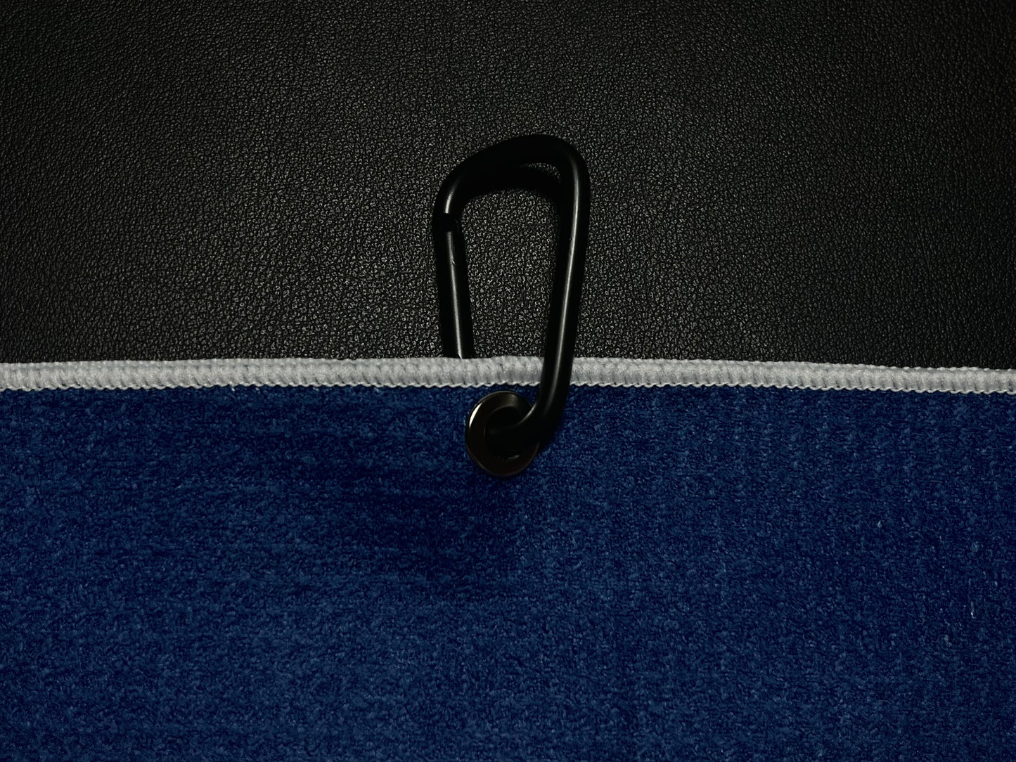 Close up of a black carabiner clipped through the grommet of a blue microfiber golf towel against a black golf bag.
