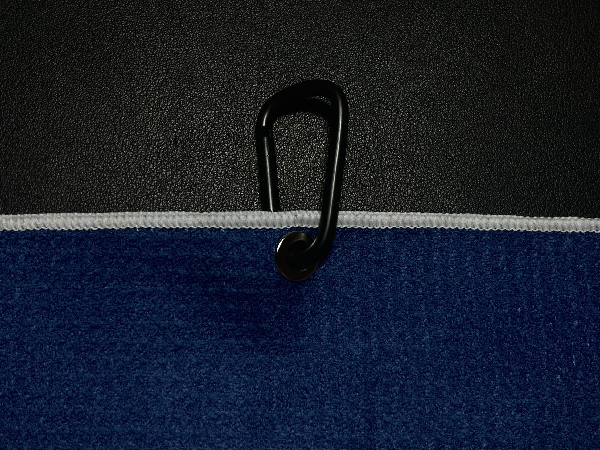 Close up of a black carabiner clipped through the grommet of a blue microfiber golf towel against a black golf bag.