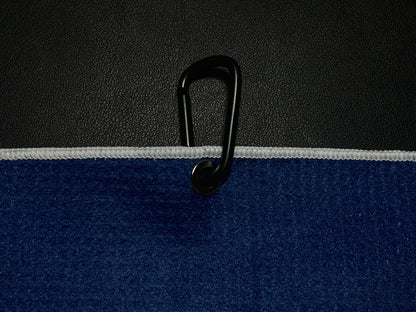 Close up of a black carabiner clipped through the grommet of a blue microfiber golf towel against a black golf bag.
