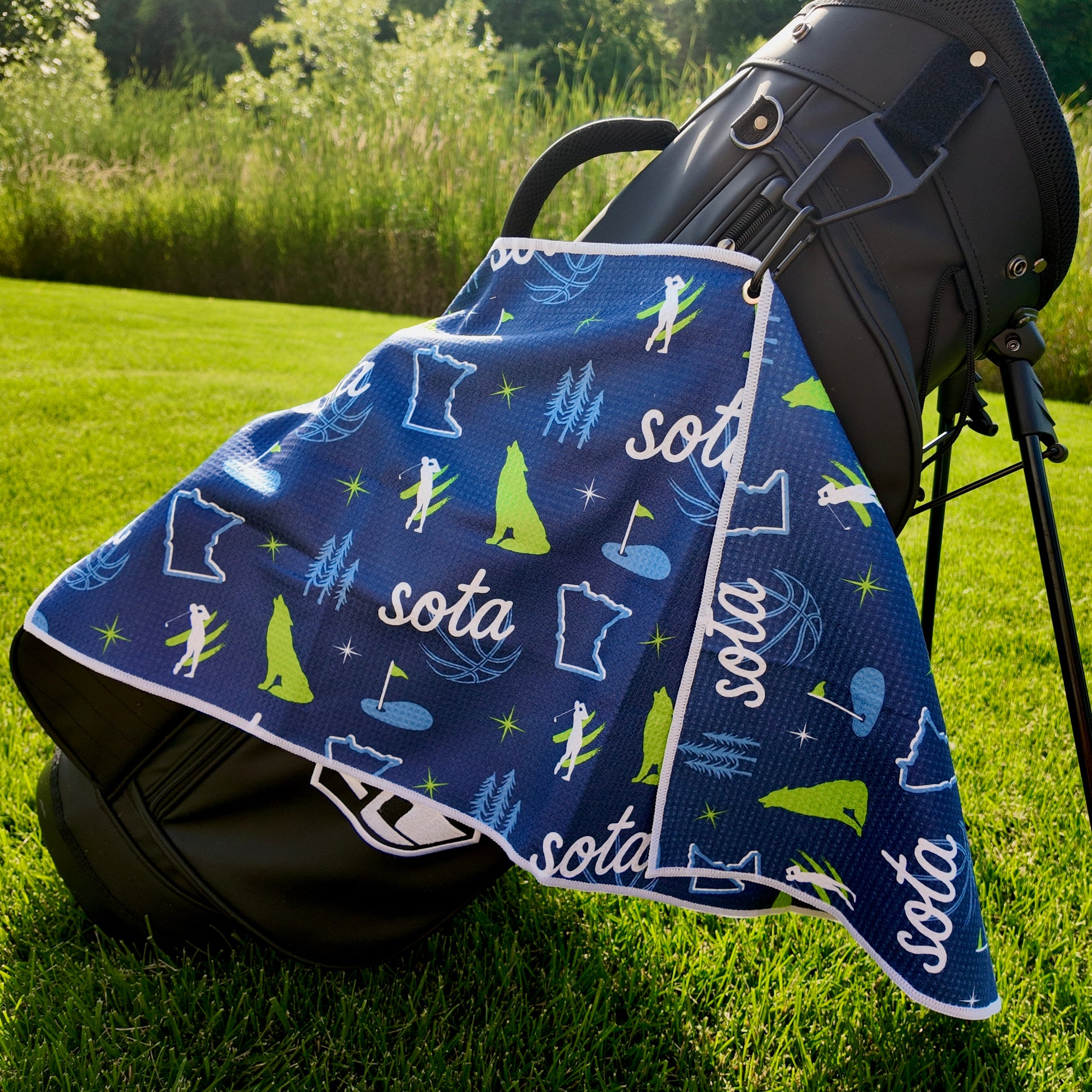 Blue and green SOTA Minnesota golf towel hanging from a black golf bag with Minnesota outlines, pine trees, golfer icons, and golf flags.