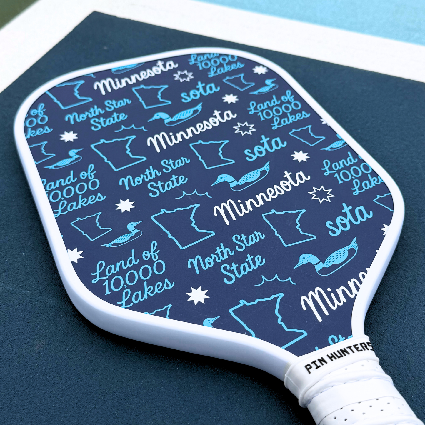 Blue Minnesota pickleball paddle close-up with white edge, light blue loons, state outlines, stars, and Land of 10,000 Lakes text.