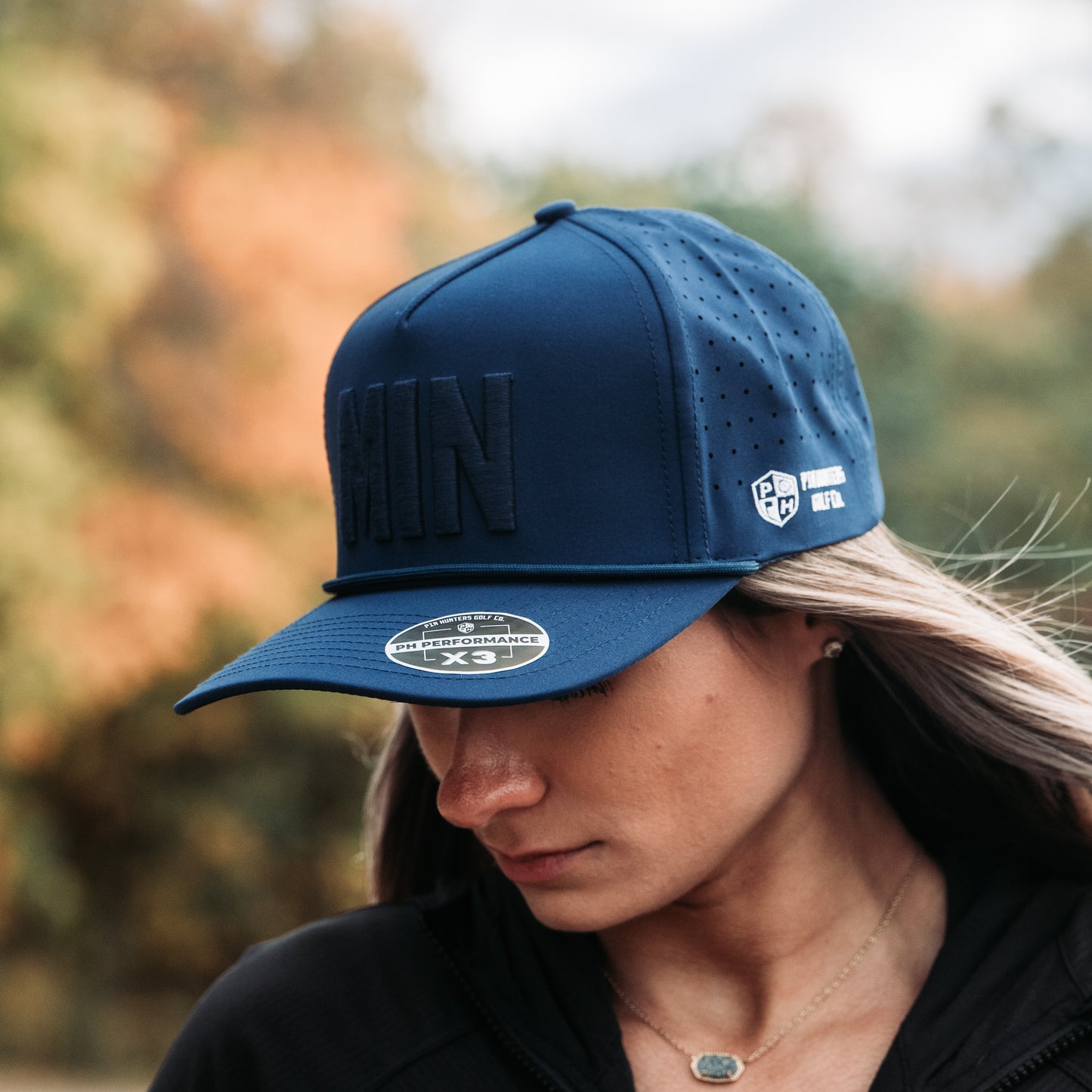 Blue tonal MIN performance golf hat on model with raised matching embroidery, side logo, perforated panels, rope detail, and PH Performance X3 sticker.