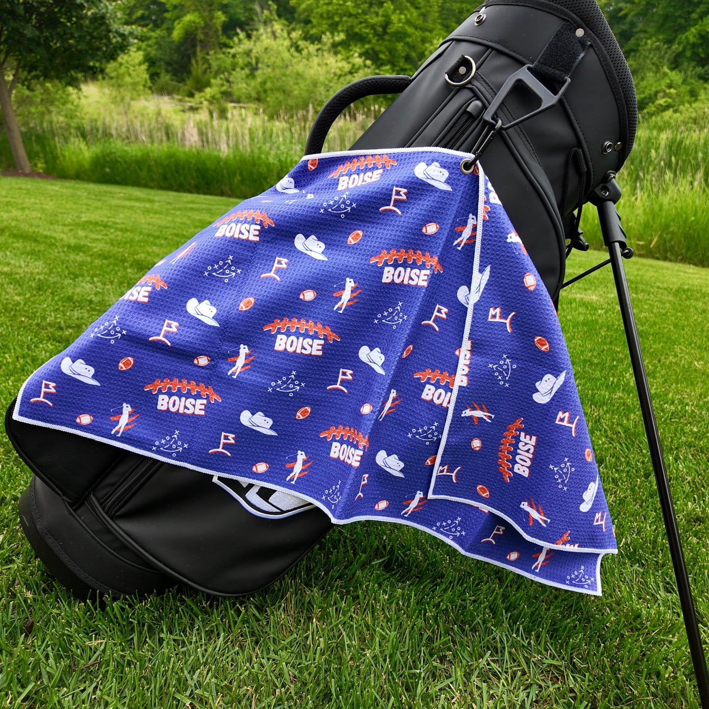 Boise State Broncos golf towel hanging on a black golf bag with blue and orange Boise text, footballs, cowboy hats, golfers, golf flags, and playbook icons.