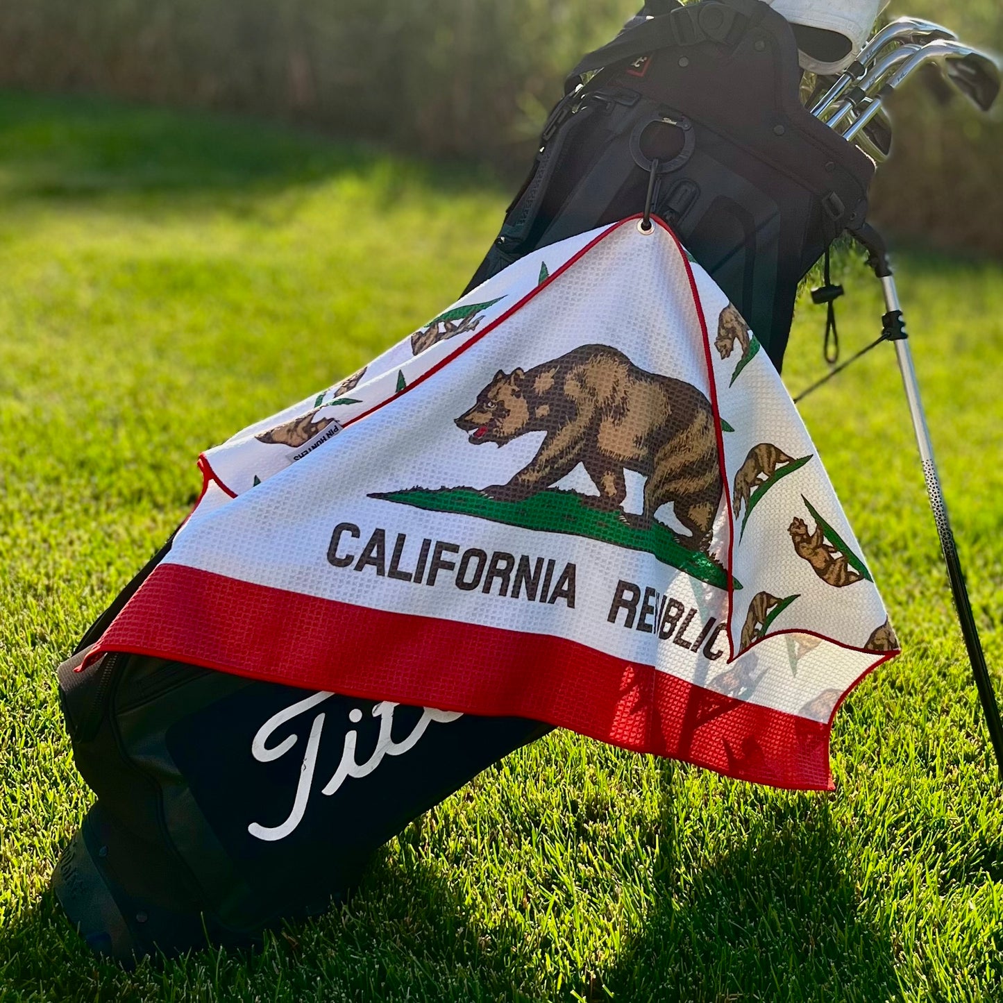 California Republic golf towel hanging from a golf bag with bear flag inspired front design, red trim, and white microfiber fabric.