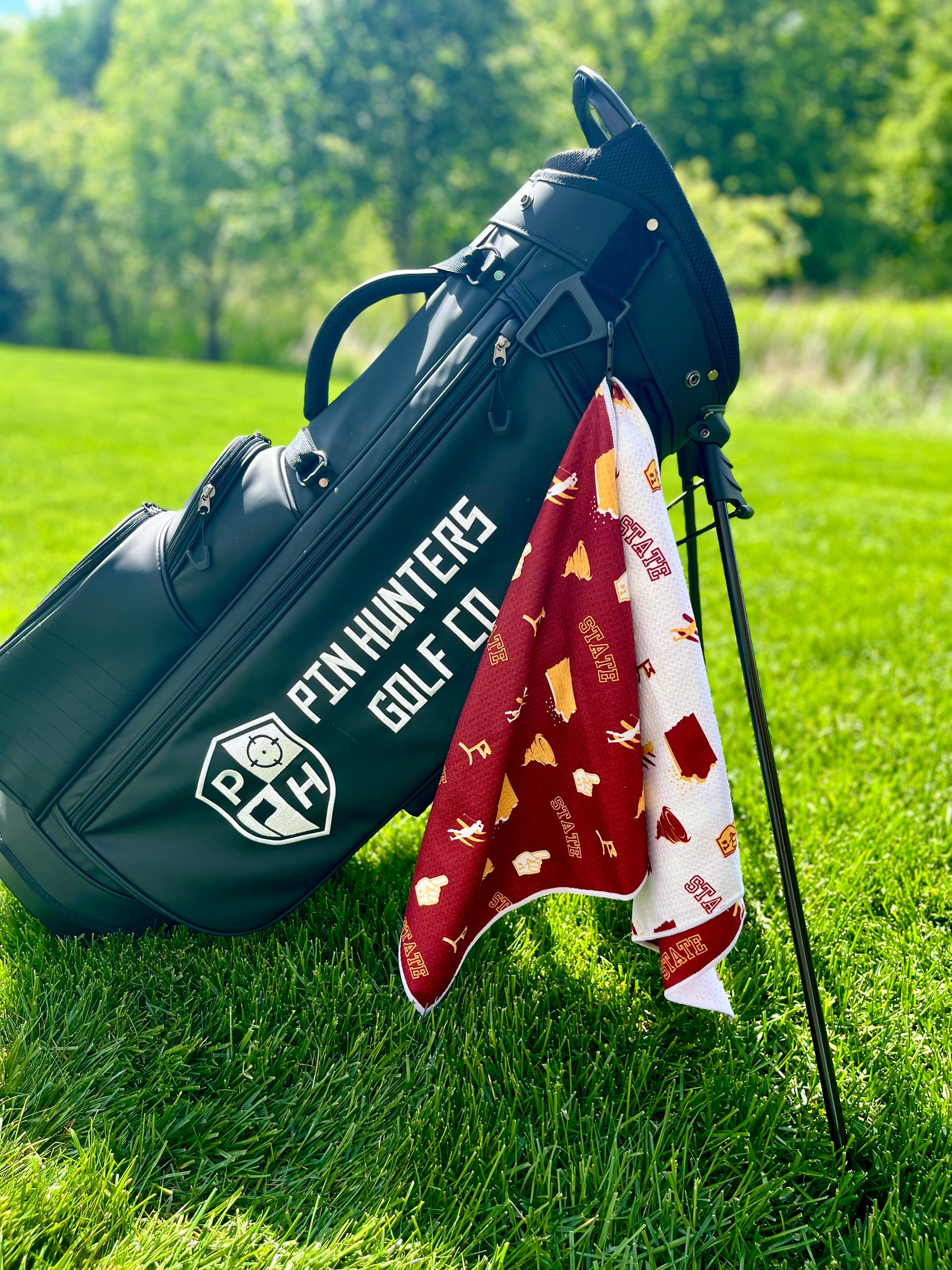 Cardinal, gold, and white Iowa State inspired golf towel hanging from a black Pin Hunters golf bag on grass.