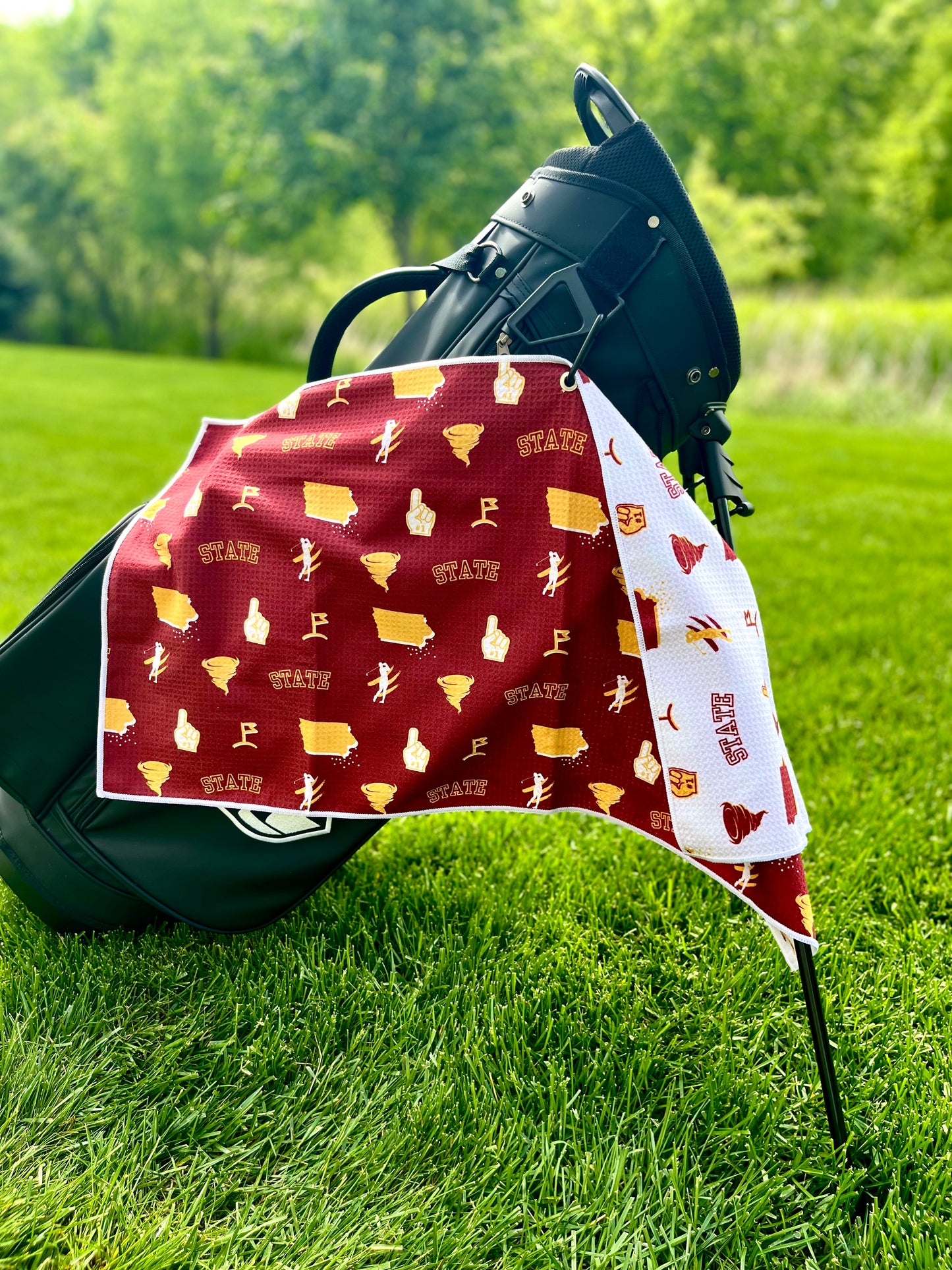 Cardinal and gold Iowa State inspired golf towel hanging from a black golf bag with state, cyclone, foam finger, golfer, and golf icons.