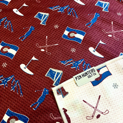 Close up of Colorado Avalanche golf towel with burgundy, blue, and white Colorado flag, mountains, hockey sticks, golfer, and golf flag graphics.