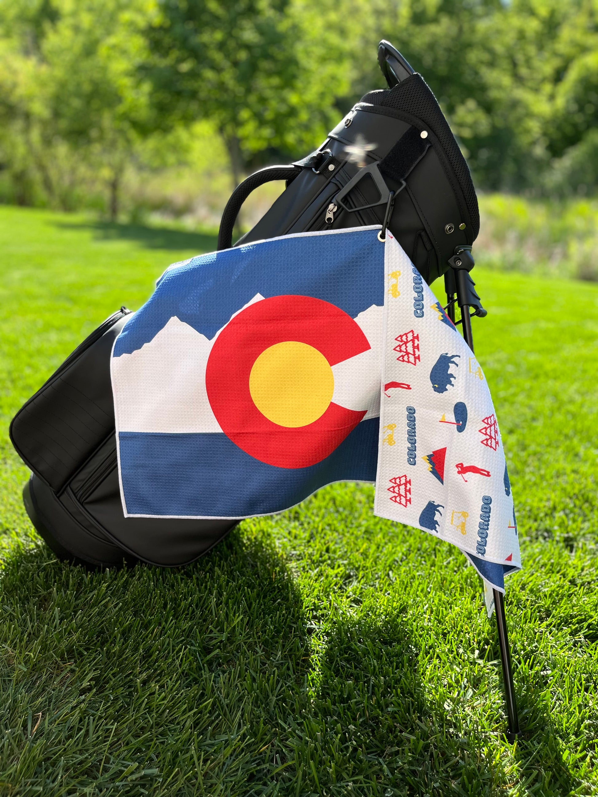 Colorado flag golf towel hanging from a black golf bag with the red C, yellow center, white mountain stripe, and blue Colorado flag front design.