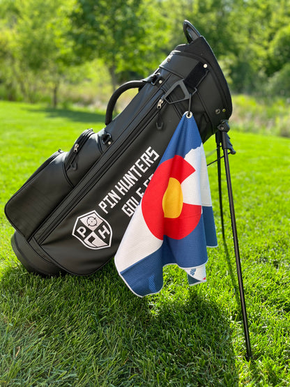 Colorado flag golf towel hanging from a black Pin Hunters golf bag, showing the blue, white, red, and yellow Colorado flag front design.