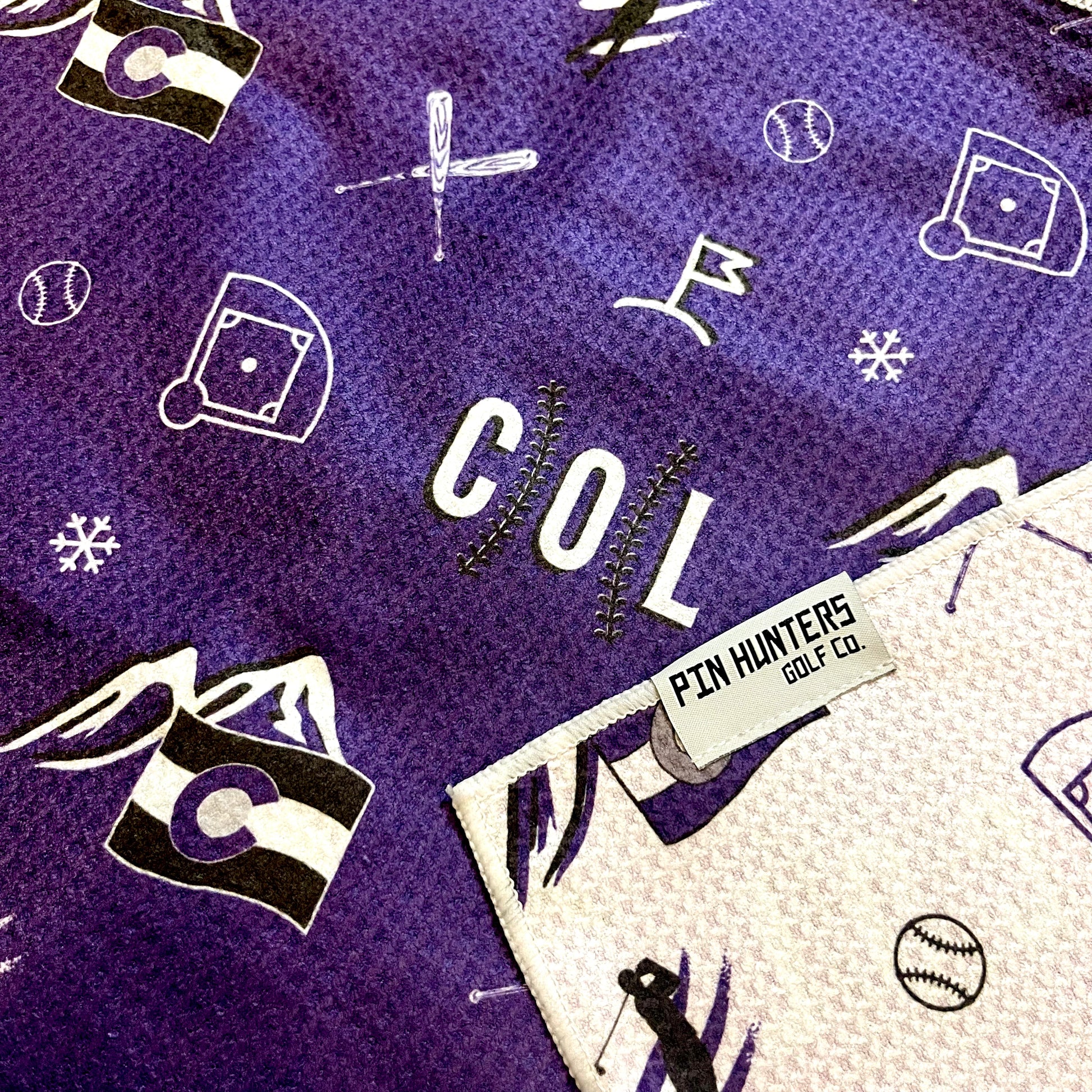 Close up of Colorado Rockies golf towel with purple, black, and white COL, mountain, baseball, golfer, and golf flag graphics.
