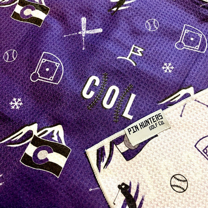 Close up of Colorado Rockies golf towel with purple, black, and white COL, mountain, baseball, golfer, and golf flag graphics.