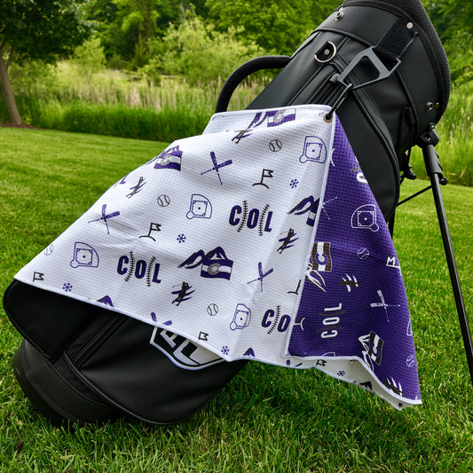 Colorado Rockies white golf towel hanging on a black golf bag with purple COL text, mountain graphics, baseballs, crossed clubs, golf flags, and purple reverse side.