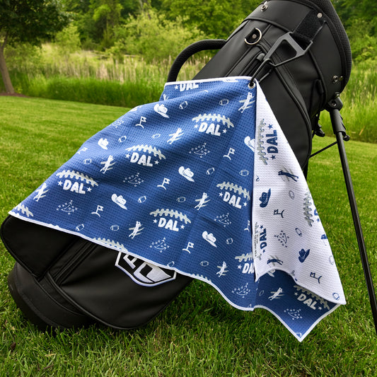 Dallas Cowboys blue golf towel hanging on a black golf bag with DAL text, white football graphics, cowboy hats, golfers, playbook, and white reverse side.