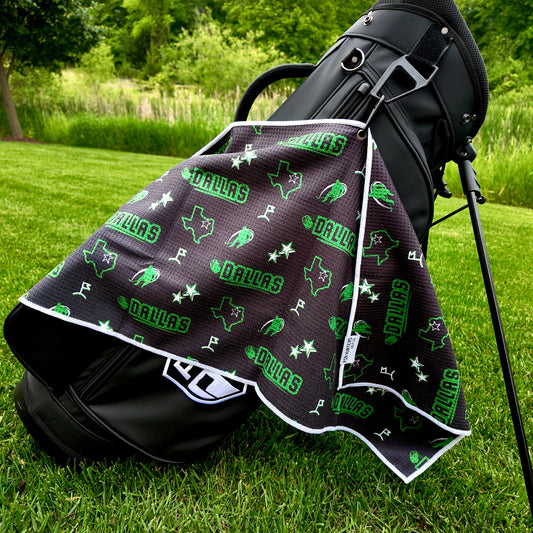 Dallas Stars black and green golf towel hanging on a black golf bag with Dallas text, Texas outlines with stars, green star graphics, golfers, and golf flags.