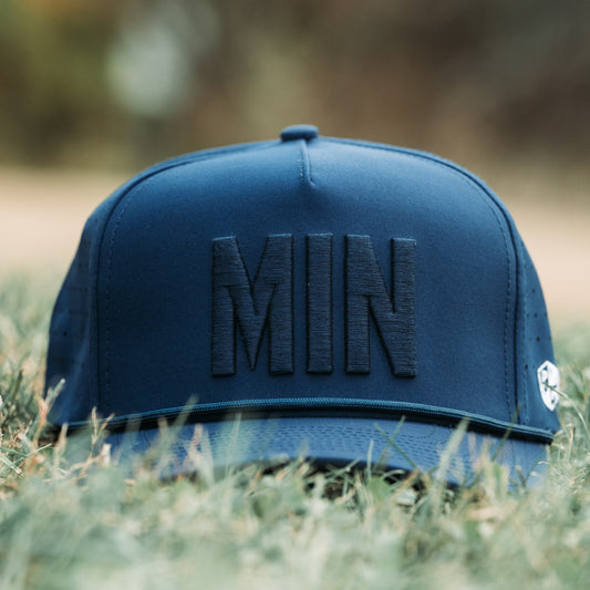Dark tonal MIN performance golf hat on grass with raised MIN embroidery, matching rope detail, flat brim, and PH Performance X3 sticker.