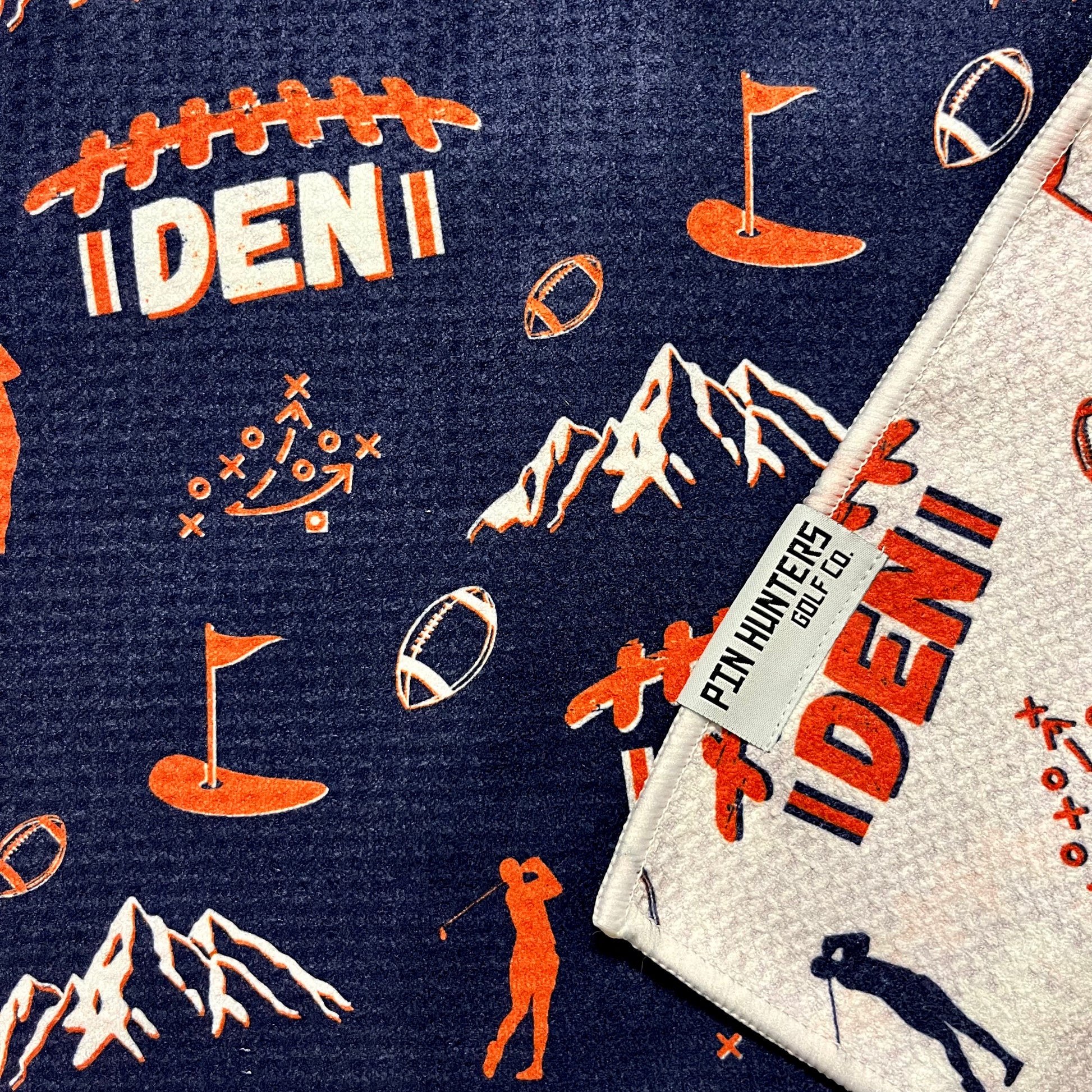 Close up of Denver Broncos golf towel with navy, orange, and white football, mountain, golfer, and flag graphics.