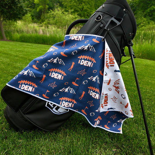 Denver Broncos navy golf towel hanging on a black golf bag with orange DEN football graphics, mountains, footballs, golfers, golf flags, and white reverse side.