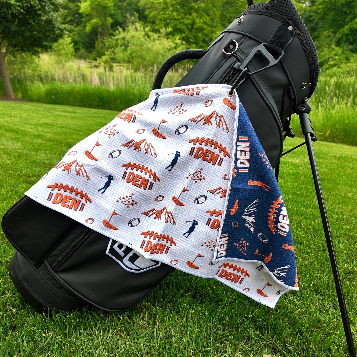 Denver Broncos white golf towel hanging on a black golf bag with orange DEN football graphics, mountains, footballs, golfers, playbook, and navy reverse side.