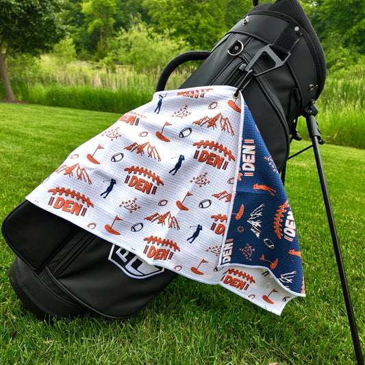 Denver Broncos white golf towel hanging on a black golf bag with orange DEN football graphics, mountains, footballs, golfers, playbook, and navy reverse side.