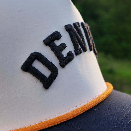 Close up of a Denver performance golf hat with black raised embroidery, white front panel, navy brim, orange rope, and stitched front seam.