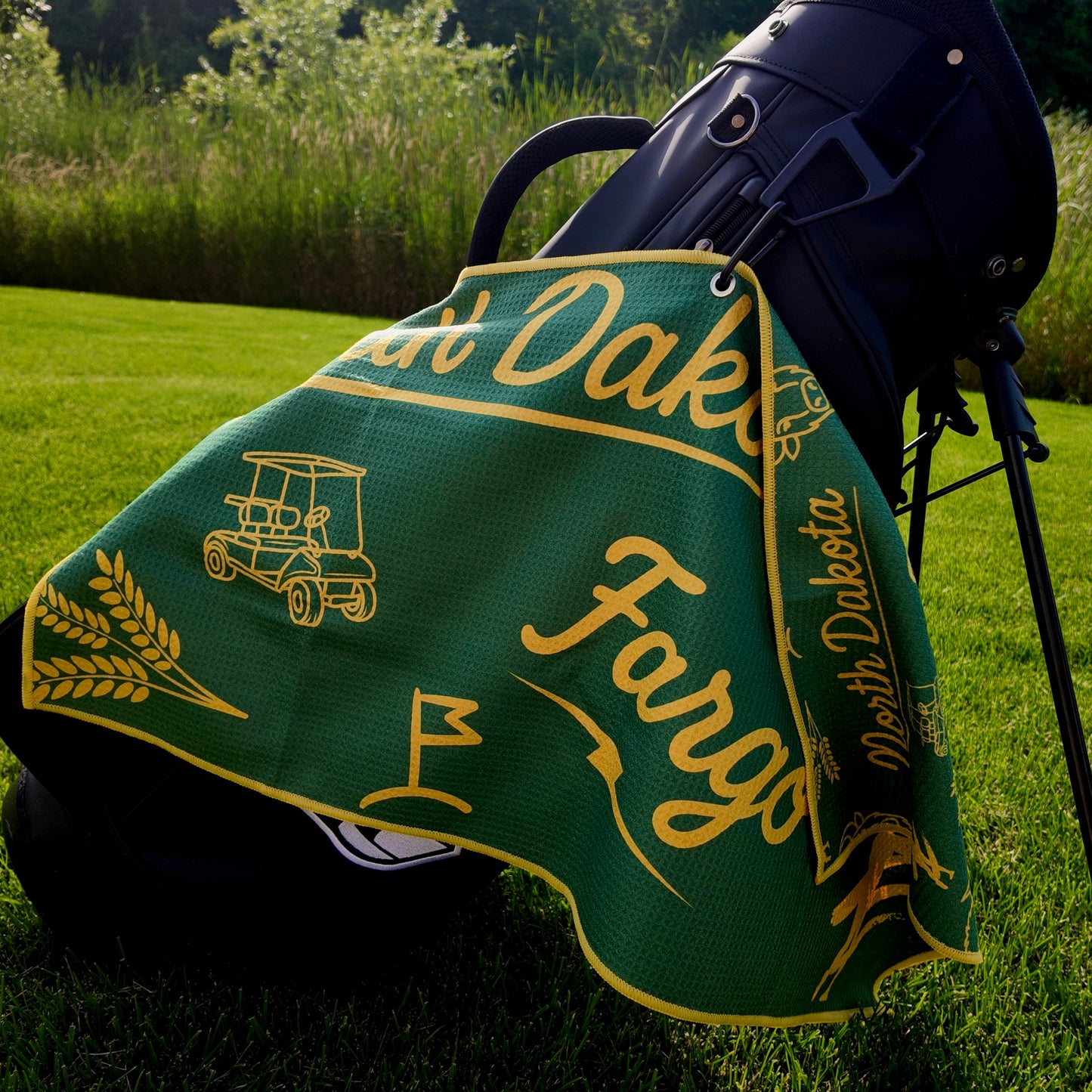 Fargo Bison NDSU golf towel hanging on a black golf bag with green and yellow Fargo script, North Dakota text, golf cart, flag, wheat, and bison graphics.