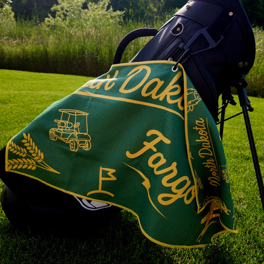 Fargo Bison NDSU golf towel hanging on a black golf bag with green and yellow Fargo script, North Dakota text, golf cart, flag, wheat, and bison graphics.