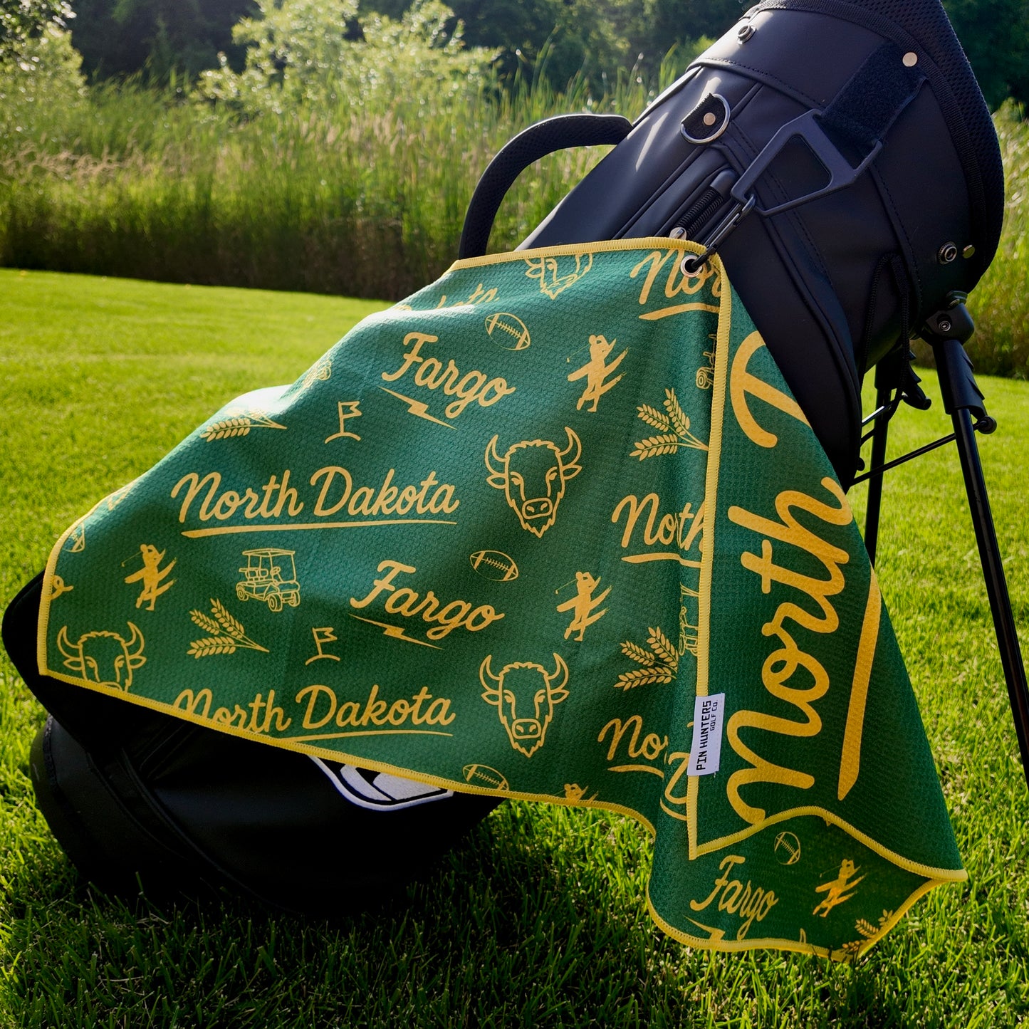 Fargo Bison NDSU golf towel hanging on a black golf bag with green and yellow North Dakota, Fargo, bison, wheat, football, golfer, golf cart, and flag graphics.