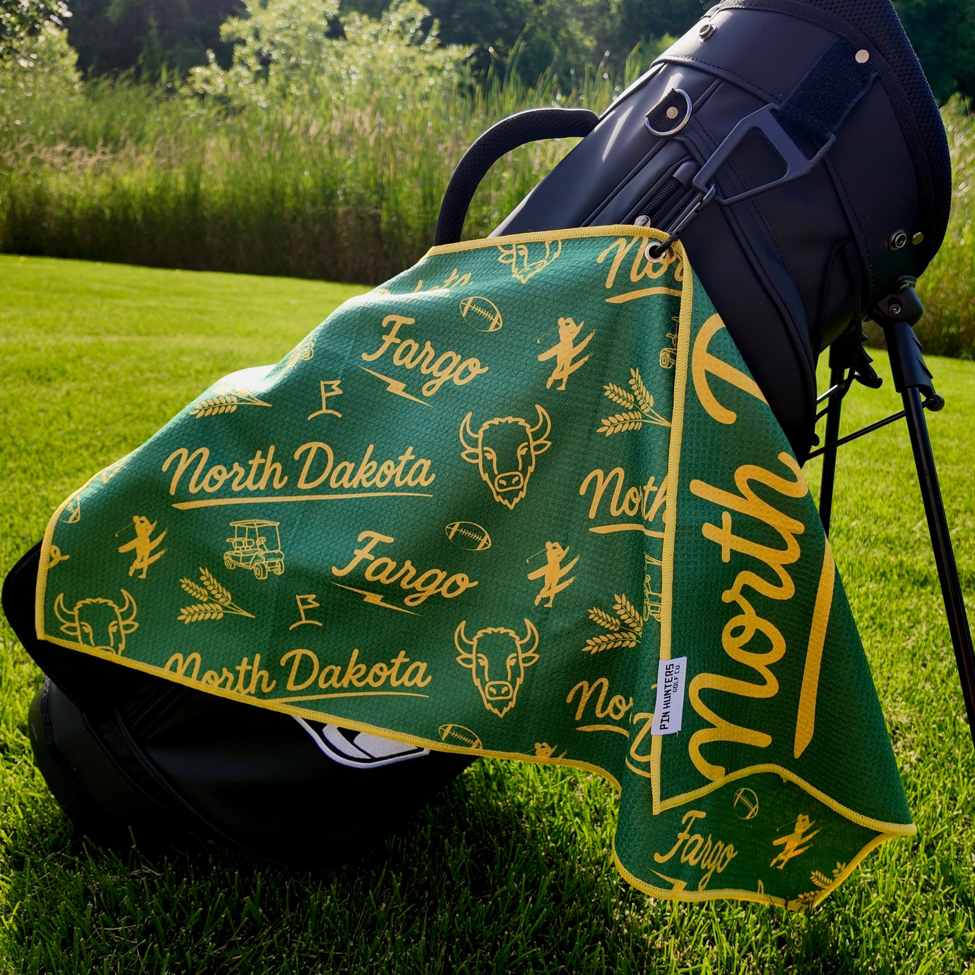 Fargo Bison NDSU golf towel hanging on a black golf bag with green and yellow North Dakota, Fargo, bison, wheat, football, golfer, golf cart, and flag graphics.