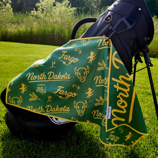 Fargo Bison NDSU golf towel hanging on a black golf bag with green and yellow North Dakota, Fargo, bison, wheat, football, golfer, golf cart, and flag graphics.