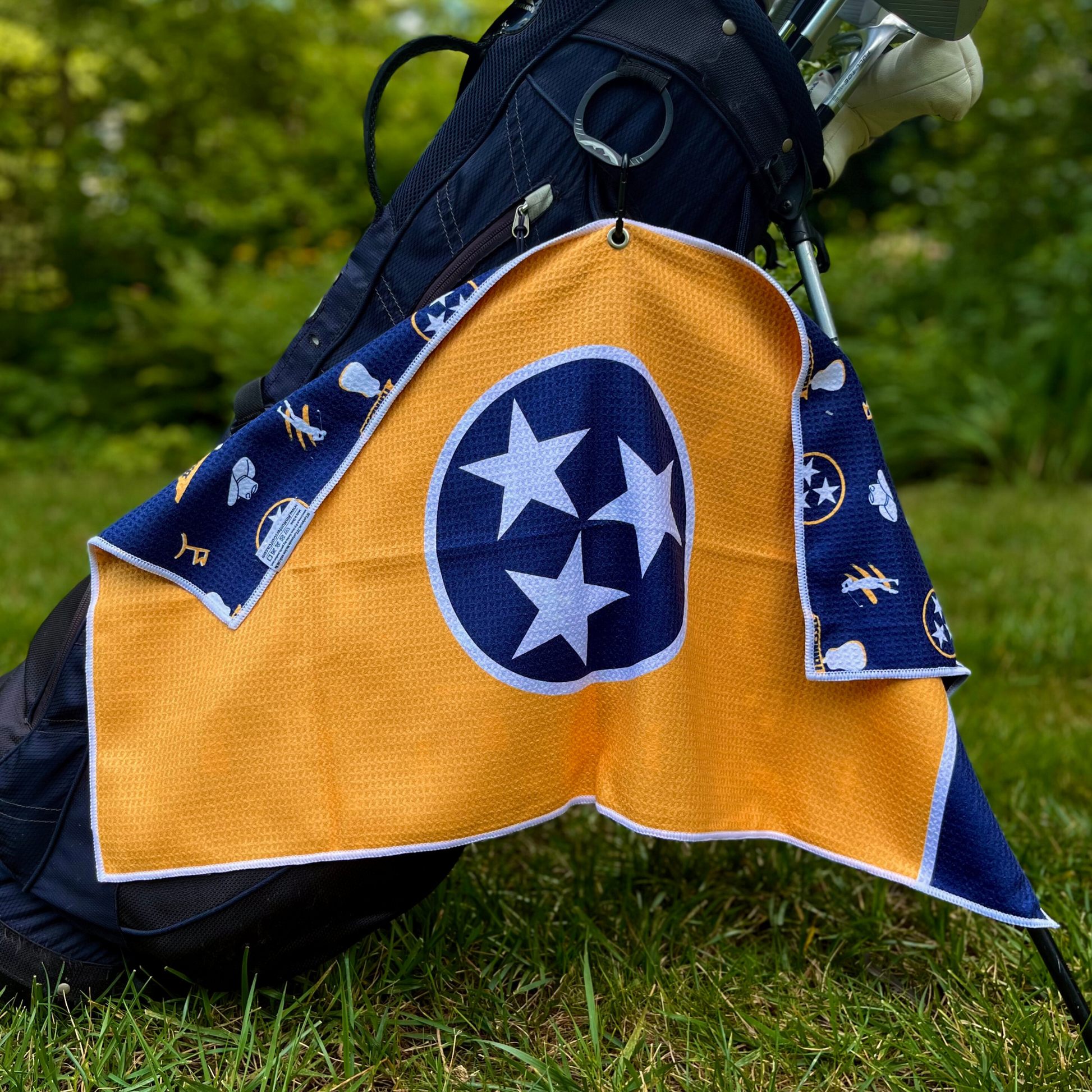 Gold Tennessee TriStar golf towel hanging from a golf bag with a large navy and white TriStar front design and navy Tennessee themed back pattern.