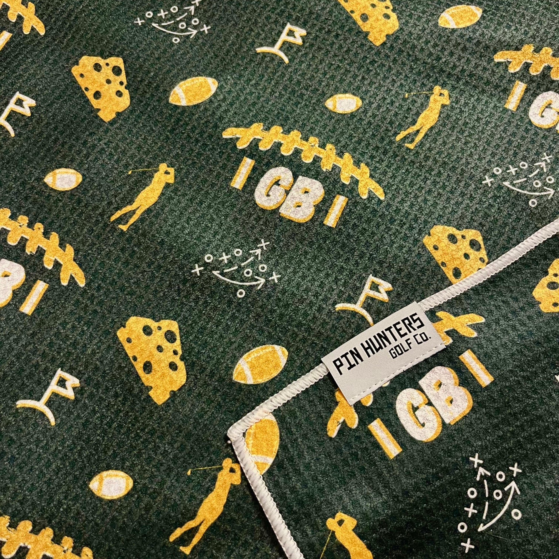 Close up of Green Bay Packers golf towel with green and yellow football, cheesehead, golfer, playbook, and golf flag graphics.
