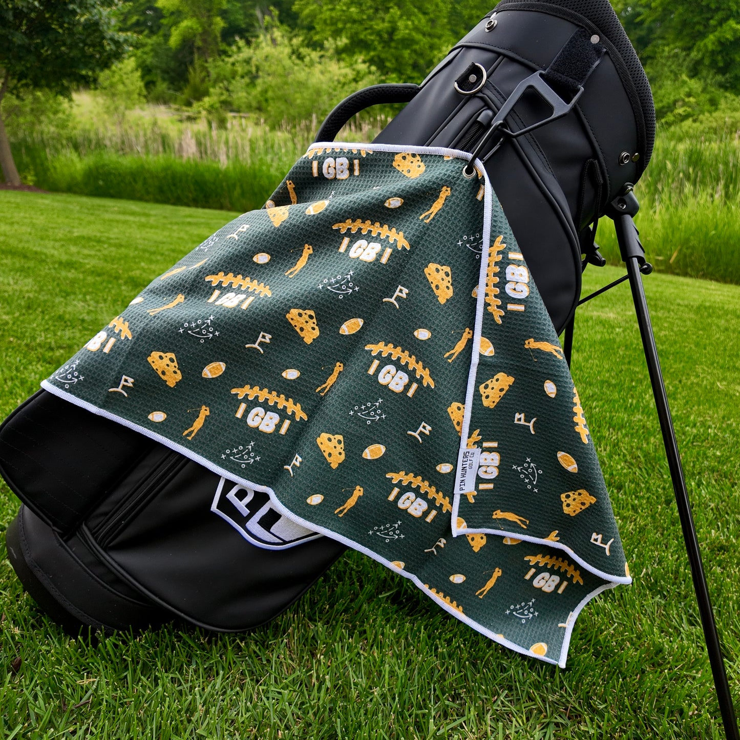 Green Bay Packers golf towel hanging on a black golf bag with green and gold footballs, cheese, golfer, playbook, golf flag, and game day golf graphics.