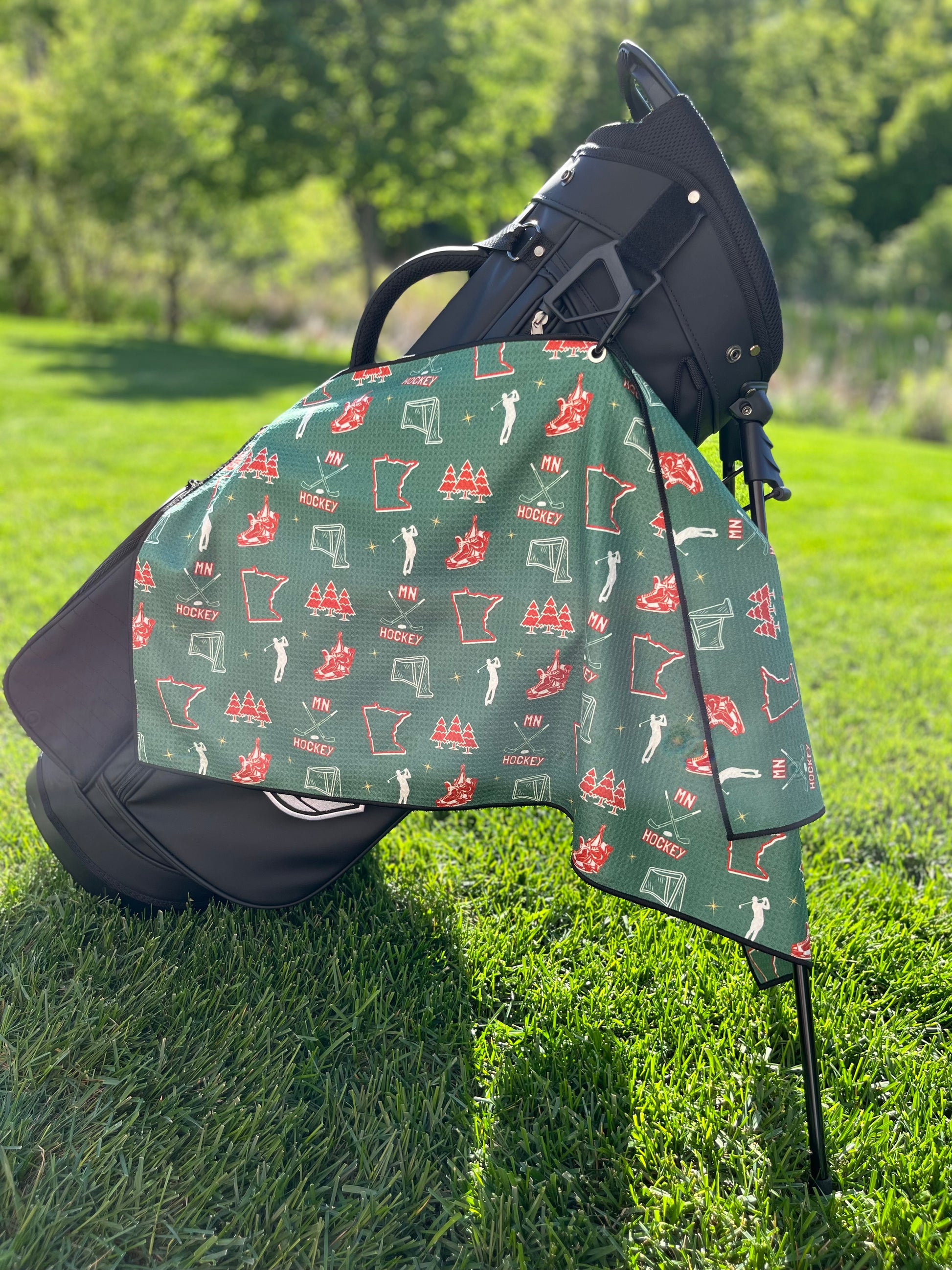 Green Minnesota Wild inspired hockey golf towel hanging from a black Pin Hunters golf bag with hockey skates, nets, MN text, Minnesota outlines, and golfer icons.