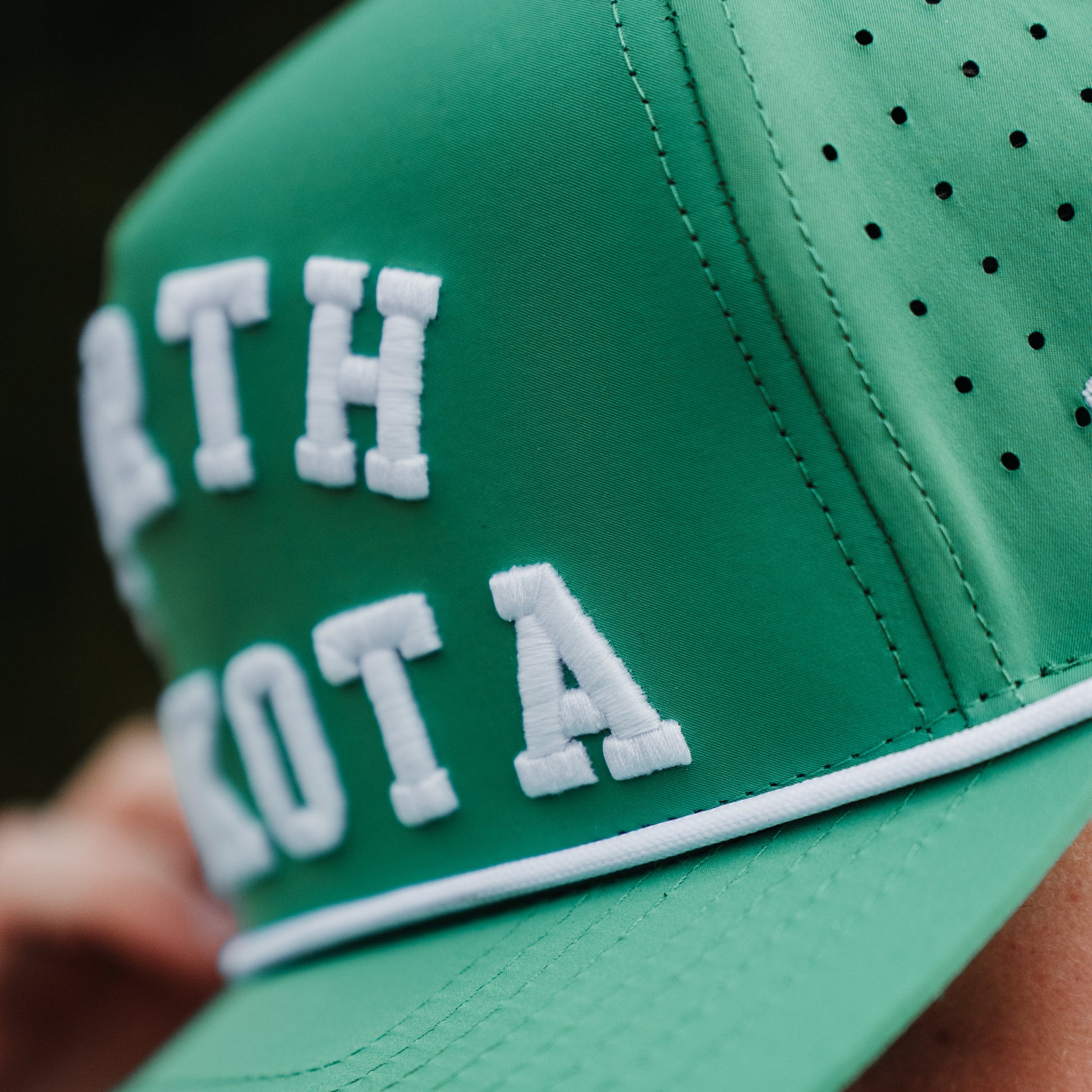 Green North Dakota performance golf hat close-up with white raised embroidery, white rope detail, and perforated side panels.