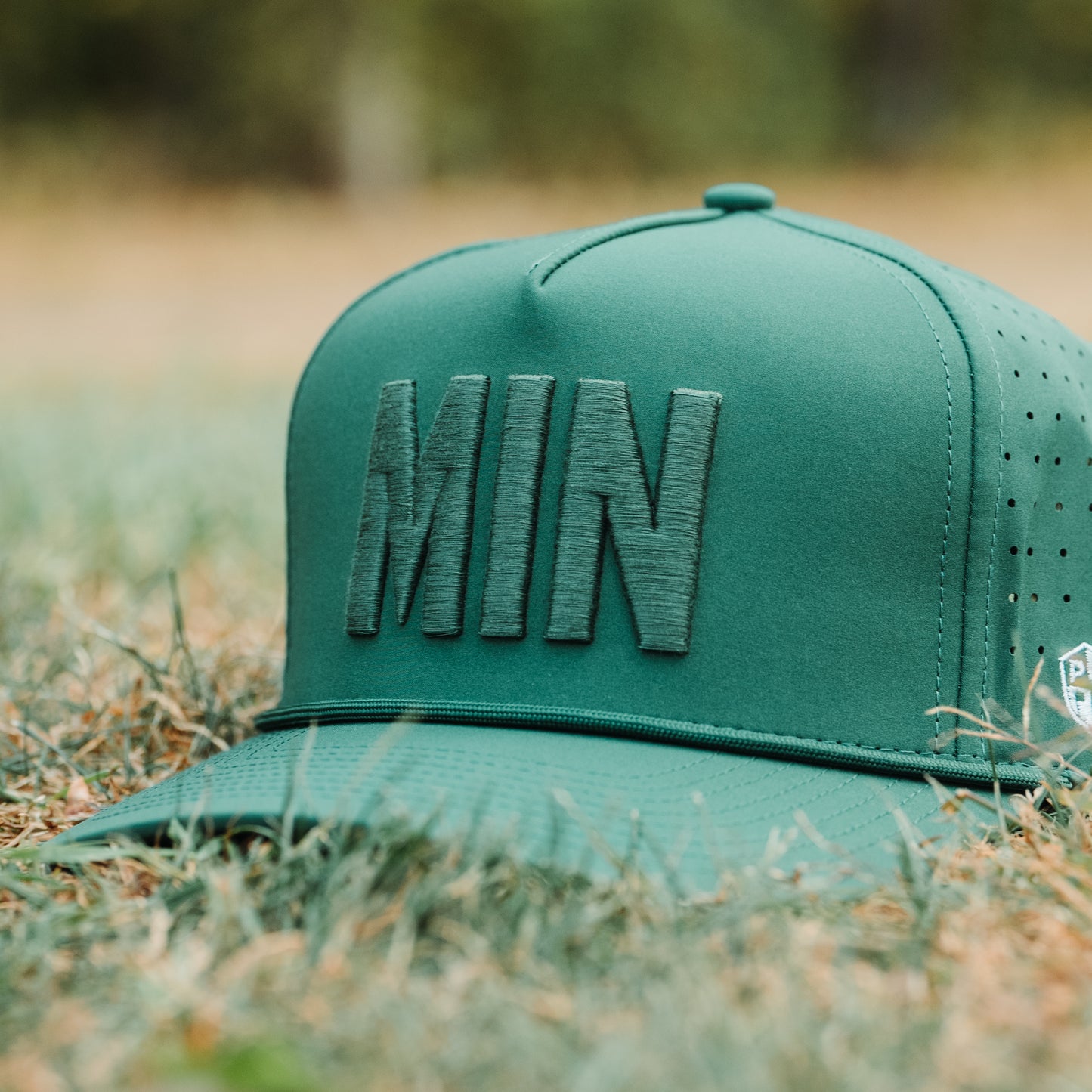 Green tonal MIN performance golf hat on grass with raised MIN embroidery, matching rope detail, flat brim, and performance fabric.