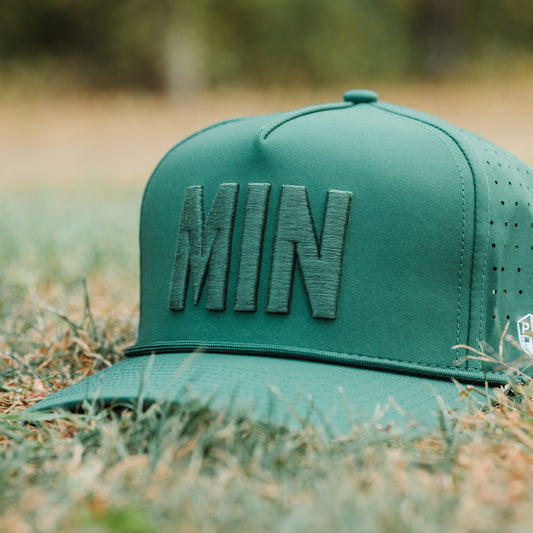 Green tonal MIN performance golf hat on grass with raised MIN embroidery, matching rope detail, flat brim, and performance fabric.