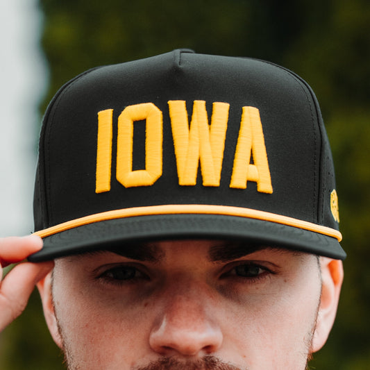 Iowa Hawkeyes black performance hat with yellow IOWA raised embroidery, yellow rope detail, perforated side panels, and lifestyle model view.
