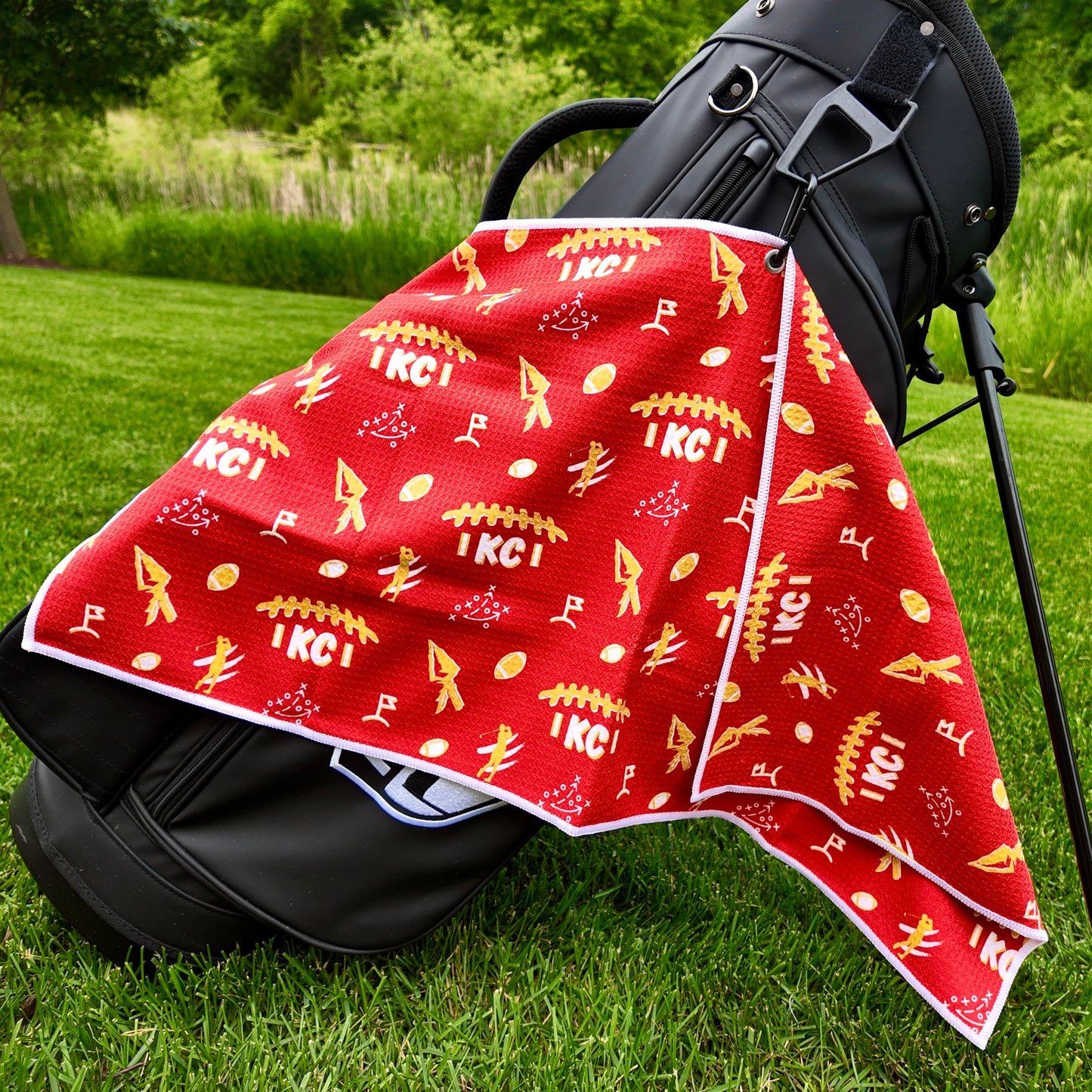 Kansas City Chiefs red golf towel hanging on a black golf bag with KC text, arrowhead graphics, footballs, golfers, golf flags, and gold and white accents.