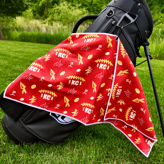 Kansas City Chiefs red golf towel hanging on a black golf bag with KC text, arrowhead graphics, footballs, golfers, golf flags, and gold and white accents.