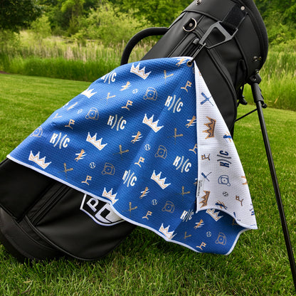 Kansas City Royals blue golf towel hanging on a black golf bag with KC text, crown graphics, baseballs, golfers, golf flags, and white reverse side.