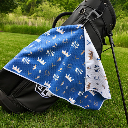 Kansas City Royals blue golf towel hanging on a black golf bag with KC text, crown graphics, baseballs, golfers, golf flags, and white reverse side.