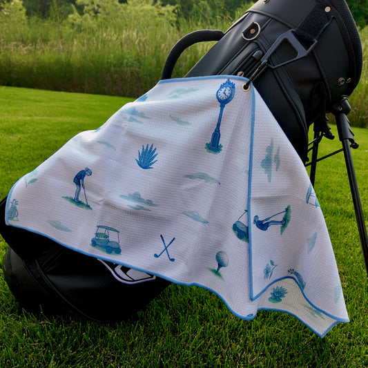 Ladies light blue golf towel hanging on a black golf bag with soft blue golf graphics, golfers, golf carts, golf balls, crossed clubs, flowers, and course icons.