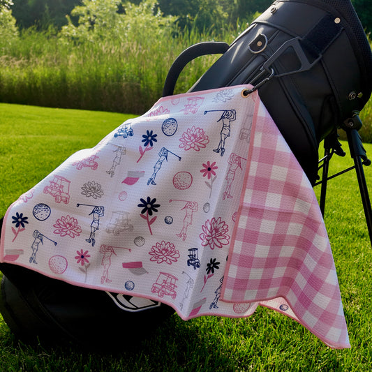 Ladies pink floral golf towel hanging on a black golf bag with golfers, flowers, golf carts, golf balls, and pink gingham reverse side.