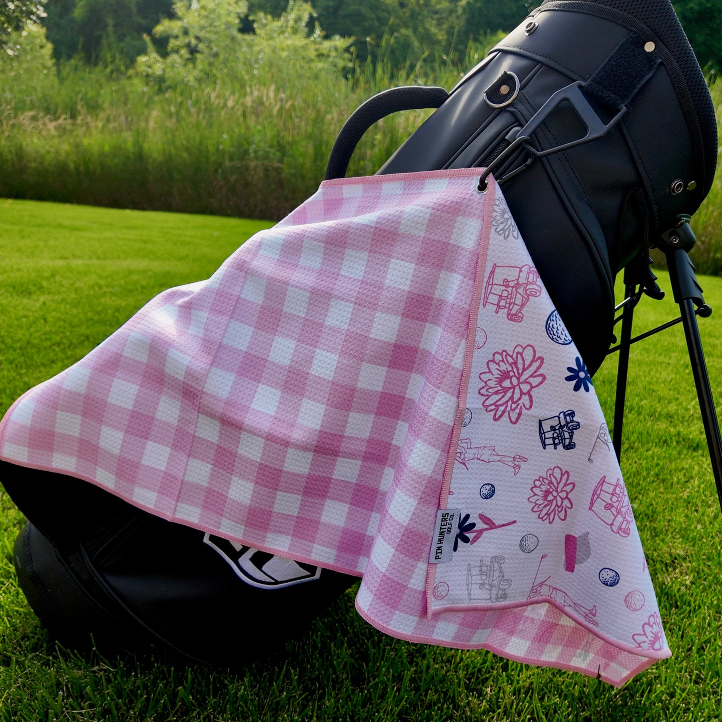 Ladies pink gingham golf towel hanging on a black golf bag with pink check pattern, floral golf graphics, golfers, golf carts, golf balls, and feminine golf design.