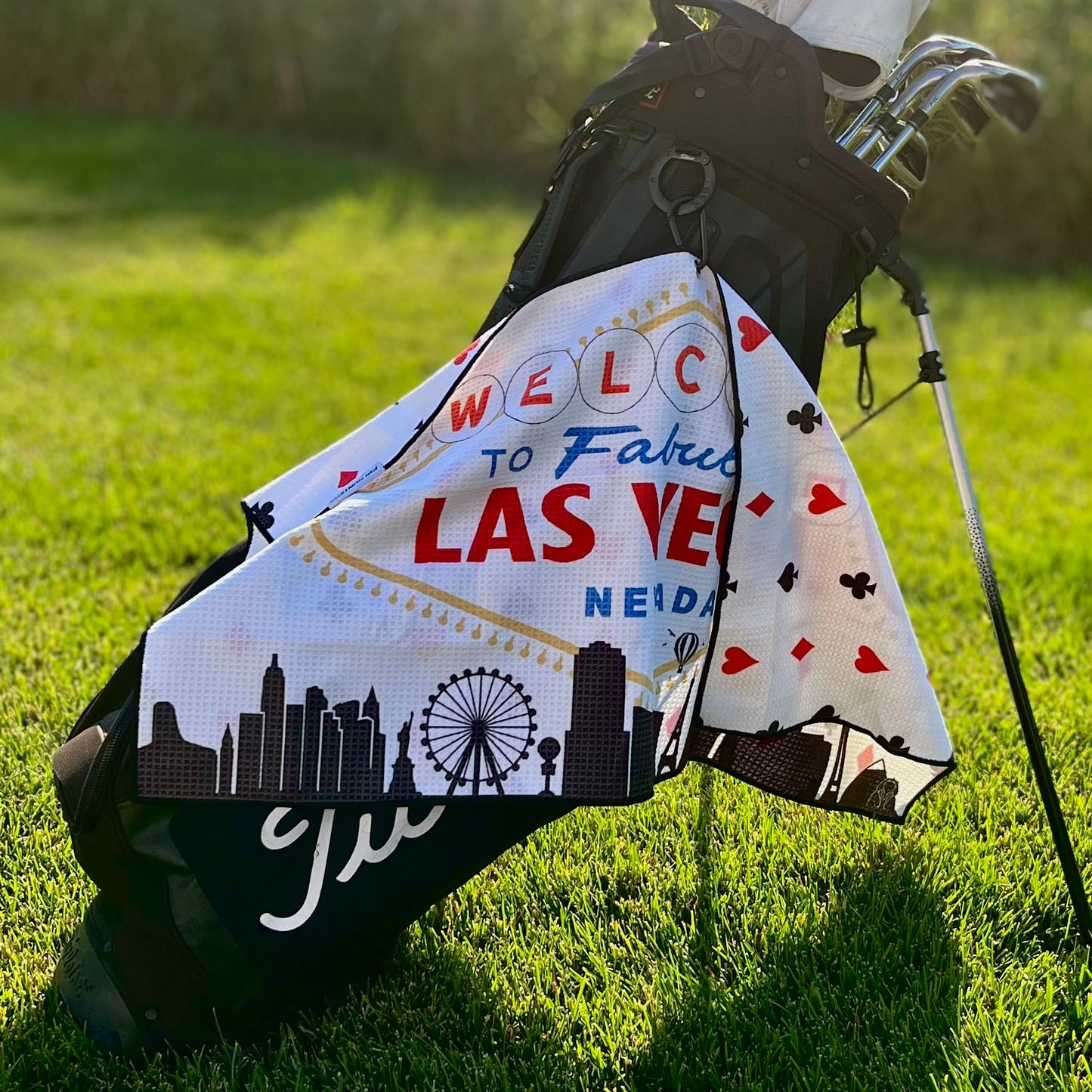 Las Vegas golf towel hanging from a black golf bag, showing the Welcome to Fabulous Las Vegas sign, skyline, ferris wheel, and playing card suit back pattern.