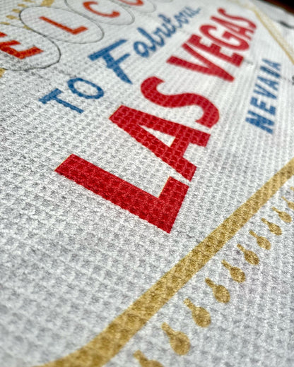 Close up of a Las Vegas golf towel showing the Welcome to Fabulous Las Vegas design, red lettering, gold sign detail, casino card icons, and waffle microfiber texture.