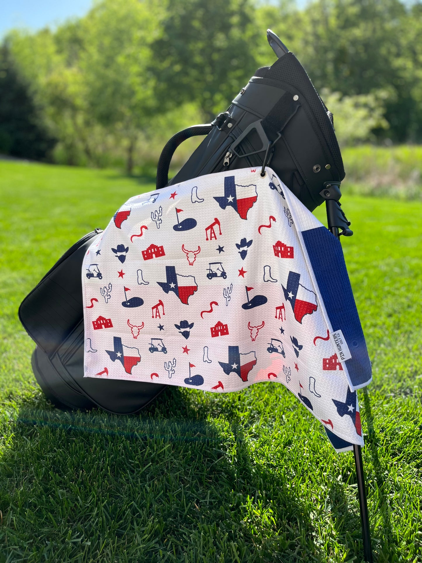 Lone Star Texas flag themed golf towel hanging from a black golf bag with red and blue Texas state, boot, longhorn, oil, cactus, Alamo, and golf icons.