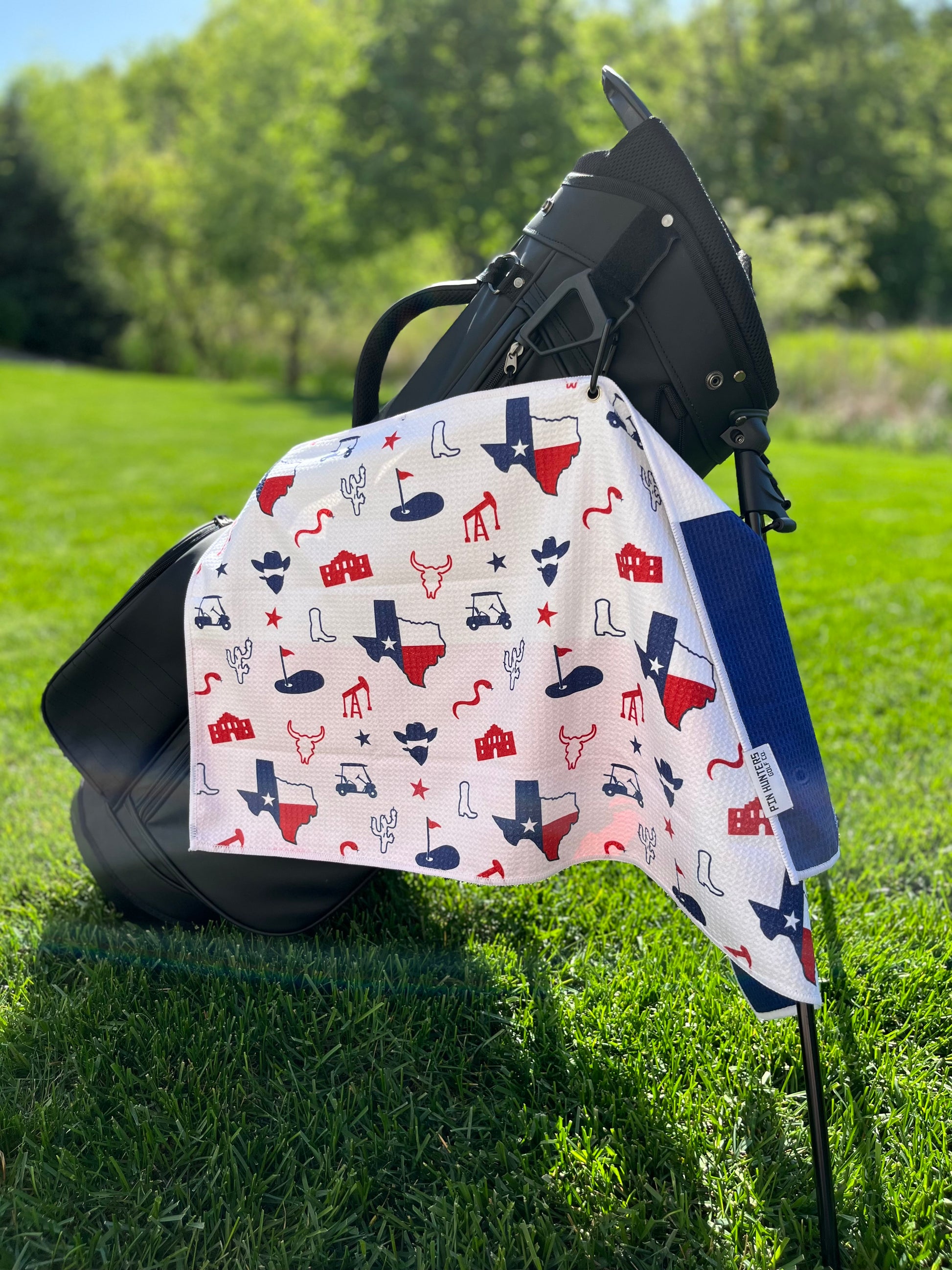 Lone Star Texas flag themed golf towel hanging from a black golf bag with red and blue Texas state, boot, longhorn, oil, cactus, Alamo, and golf icons.