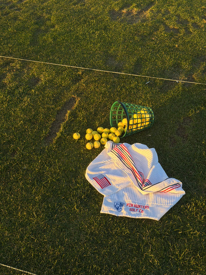 Pin Hunters Looper cotton golf towels on grass near a green range basket and yellow golf balls with USA flag embroidery and striped towel details.