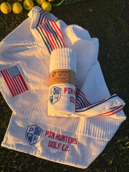 Pin Hunters Looper cotton golf towels on grass with USA flag embroidery, ribbed white towel texture, striped towel detail, and packaged rolled towel.